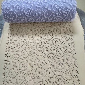 May include: A lavender clay roller with a repeating pattern of cat faces, hearts, and paw prints. The roller is pressed into a piece of clay, creating an embossed design of the same pattern. The clay is a light beige color.
