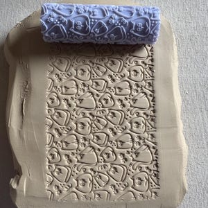 May include: A light purple clay roller with a repeating pattern of cartoon cat faces and paw prints, resting on a piece of beige clay. The roller has been used to imprint the cat face design into the clay, creating a textured surface.