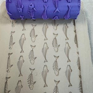 May include: A purple cylindrical rolling pin with a raised fish skeleton pattern, shown pressing a repeating fish bone design into a flat, light gray slab of clay. The tool features a continuous texture for creating impressions in soft materials.