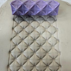 May include: A cylindrical purple clay texture roller with a raised diamond grid pattern positioned above a flat, light gray slab of clay that has been impressed with the same repeating diamond pattern, creating a deep, uniform geometric texture.