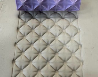 May include: A cylindrical purple clay texture roller with a raised diamond grid pattern positioned above a flat, light gray slab of clay that has been impressed with the same repeating diamond pattern, creating a deep, uniform geometric texture.