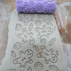 May include: A light purple cylindrical rolling pin with a raised pattern of gingerbread people, snowflakes, and holly leaves. The rolling pin is positioned at the top of a flat, light gray slab of clay that has been embossed with the same design.