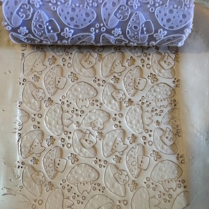 May include: A clear rolling pin with a mushroom pattern is pressed into a light-colored clay surface. The pattern includes cartoon mushrooms and small flowers. The rolling pin is made of a clear material, and the clay is a neutral color.
