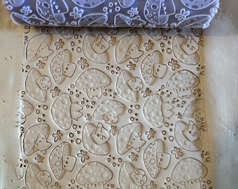 May include: A clear rolling pin with a mushroom pattern is pressed into a light-colored clay surface. The pattern includes cartoon mushrooms and small flowers. The rolling pin is made of a clear material, and the clay is a neutral color.