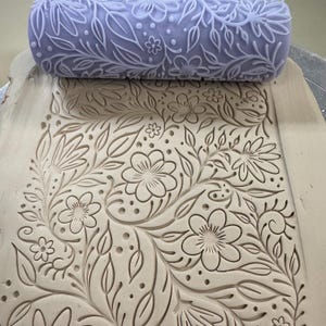 May include: A light purple clay roller with a floral design is pressed onto a piece of clay, creating an embossed pattern. The design includes flowers, leaves, and dots. The clay is a light beige color.