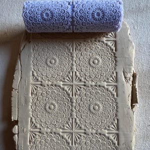 May include: A light purple clay roller with a floral pattern rests above a piece of clay imprinted with the same design. The clay has a rectangular shape with the floral pattern repeating in a grid. The roller is made of a smooth material.