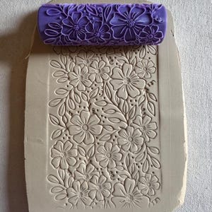 May include: A purple floral pattern rolling pin rests on a clay slab with an embossed floral design. The rolling pin has a repeating pattern of flowers and leaves. The clay slab has a similar embossed floral design.