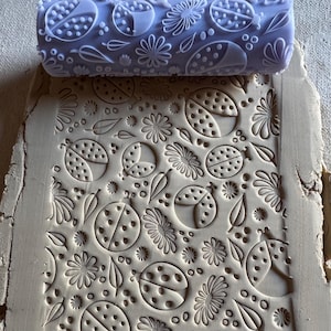 May include: A light purple rolling pin with a ladybug and floral pattern is shown next to a piece of clay with the same embossed design. The clay is beige and the rolling pin is made of a smooth material. The design includes ladybugs, flowers, and leaves.