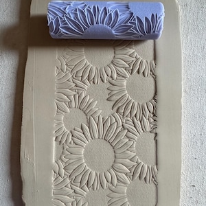 May include: A light purple clay roller with a sunflower design rests above a clay slab with an embossed sunflower pattern. The roller is cylindrical, and the sunflowers are detailed. The clay slab is a light beige color.