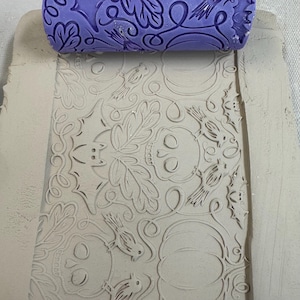 May include: A purple cylindrical clay texture roller with a repeating pattern of skulls, bats, birds, pumpkins, and leaves. The roller is positioned at the top of a rectangular slab of light gray clay, which shows the embossed impression of the design.