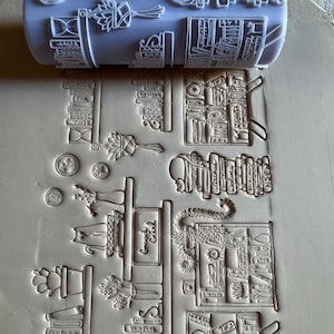 May include: A white, cylindrical rolling stamp with a detailed design of bookshelves, plants, and decorative elements. The stamp has various text elements, including "Fantasy," "Vampires," and "Witches." The stamped impression is on a light beige surface.