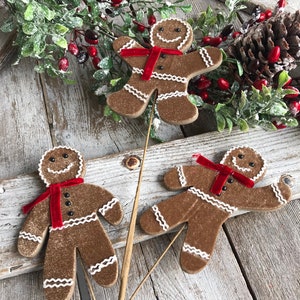 May include: Three gingerbread man ornaments with white icing trim and red velvet scarves. The brown gingerbread men have black button eyes and are arranged on a rustic wooden surface with holiday greenery.