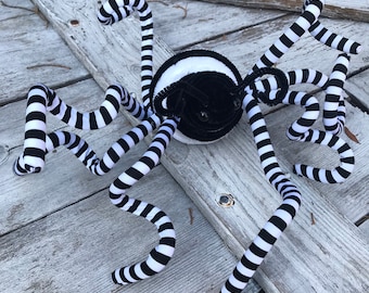 May include: A plush spider decoration with a black and white striped body and legs. The spider's body is black with a white stripe around the middle. The legs are made of a flexible material.