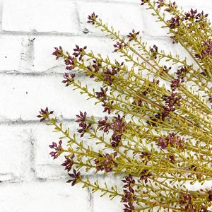 May include: A bunch of dried purple flowers with green stems against a white brick wall background.
