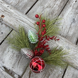 May include: A Christmas decoration featuring a green pine sprig with red berries, a silver pine cone, a green pine cone, and a red and white ornament.