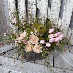 May include: A floral arrangement in a galvanized metal container. The arrangement includes peach-colored peonies, pink tulips, and greenery, with a moss-covered bunny figure. The background is a weathered, white-painted wooden surface.