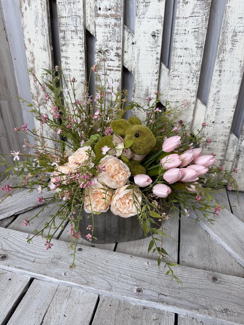 May include: A floral arrangement in a galvanized metal container. The arrangement includes peach-colored peonies, pink tulips, and greenery, with a moss-covered bunny figure. The background is a weathered, white-painted wooden surface.