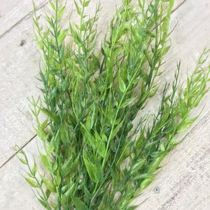 May include: A bunch of artificial green leafy stems with long, thin leaves. The stems are arranged in a fan shape and are lying on a light wood surface.