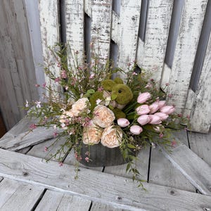 May include: A floral arrangement in a galvanized metal container. The arrangement includes pink tulips, peach-colored peonies, and a green plush bunny. Greenery and small pink flowers are interspersed throughout the arrangement. The background is a weathered white wooden surface.