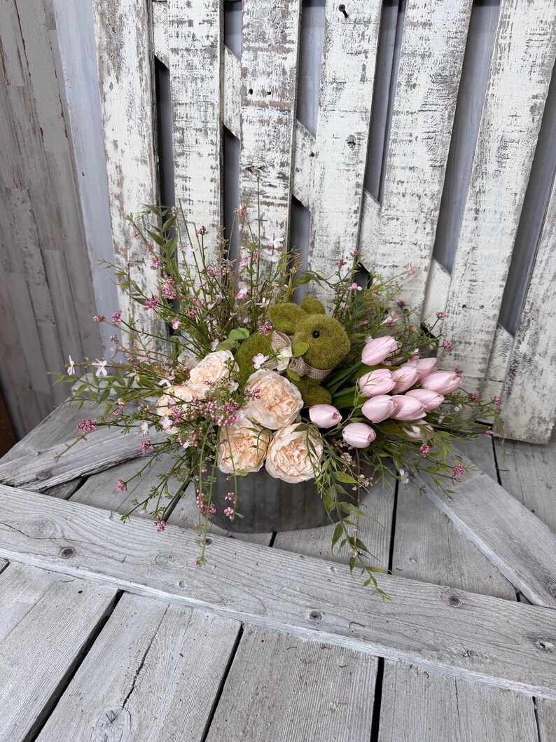 May include: A floral arrangement in a galvanized metal container. The arrangement includes pink tulips, peach-colored peonies, and a green plush bunny. Greenery and small pink flowers are interspersed throughout the arrangement. The background is a weathered white wooden surface.