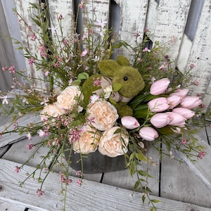 May include: An Easter-themed floral arrangement in a metal container. It features a moss-covered bunny, peach-colored peonies, pink tulips, and delicate pink and green foliage. The arrangement is set against a weathered, white wooden backdrop.