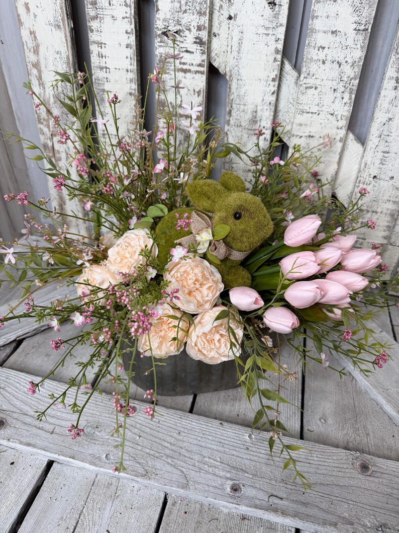 May include: An Easter-themed floral arrangement in a metal container. It features a moss-covered bunny, peach-colored peonies, pink tulips, and delicate pink and green foliage. The arrangement is set against a weathered, white wooden backdrop.