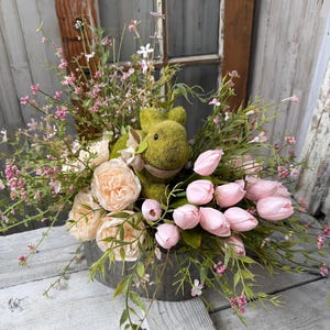 May include: A spring floral arrangement in a galvanized metal container. The arrangement features a moss-covered bunny, pink tulips, peach-colored roses, and delicate pink and green foliage. The background includes a rustic wooden window.