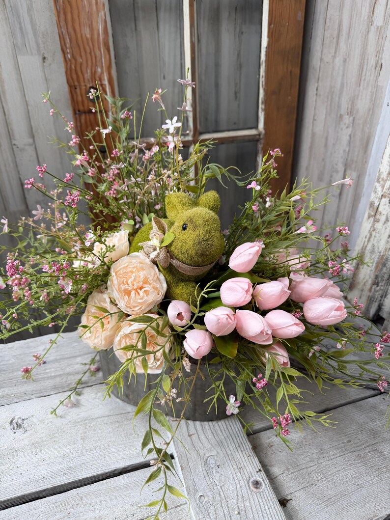May include: A spring floral arrangement in a galvanized metal container. The arrangement features a moss-covered bunny, pink tulips, peach-colored roses, and delicate pink and green foliage. The background includes a rustic wooden window.