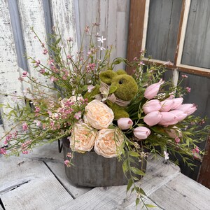May include: A floral arrangement in a galvanized metal container. It features a green bunny, pink tulips, peach roses, and various greenery and small pink flowers. The arrangement sits on a weathered wooden surface.