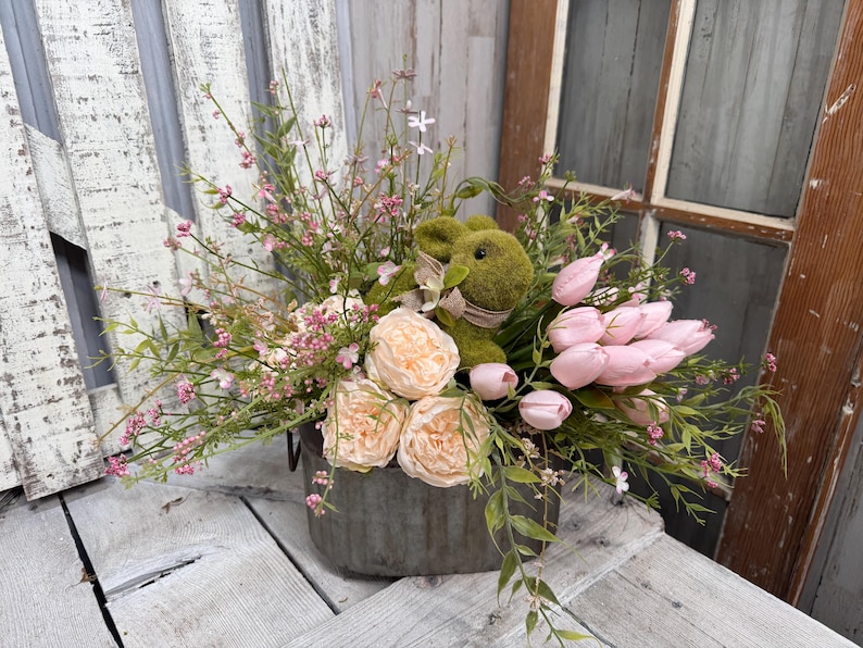 May include: A floral arrangement in a galvanized metal container. It features a green bunny, pink tulips, peach roses, and various greenery and small pink flowers. The arrangement sits on a weathered wooden surface.