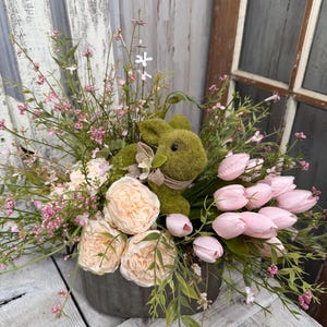 May include: A decorative Easter arrangement featuring a moss-covered bunny nestled among pink tulips, peach-colored peonies, and delicate pink and green foliage. The arrangement is in a gray metal container, perfect for spring decor.