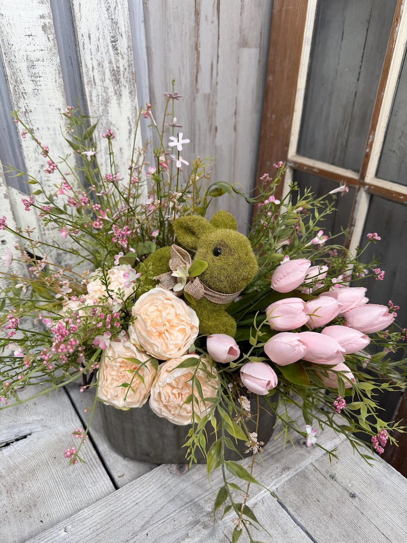 May include: A decorative Easter arrangement featuring a moss-covered bunny nestled among pink tulips, peach-colored peonies, and delicate pink and green foliage. The arrangement is in a gray metal container, perfect for spring decor.