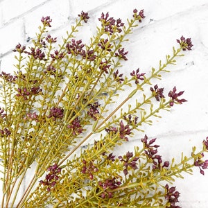 May include: A bunch of artificial greenery with small purple flower buds. The stems are green and the leaves are a light green color.