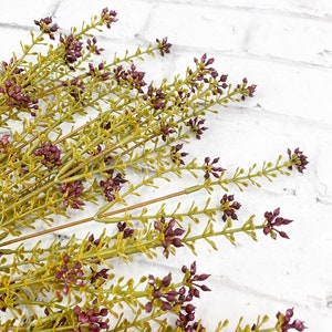 May include: A bunch of artificial greenery with small purple flowers. The stems are green and the leaves are a light green color.