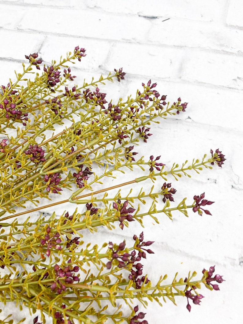 May include: A bunch of artificial greenery with small purple flowers. The stems are green and the leaves are a light green color.