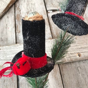 May include: Two black top hats with red bands and a small red cardinal on one hat. The hats are on a green pine branch with a bird's nest and egg on top of one hat.
