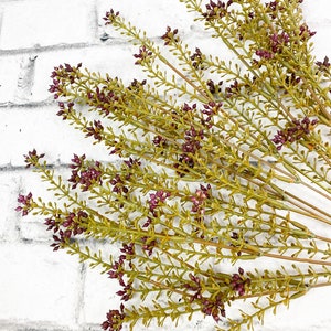 May include: A bunch of artificial greenery with small purple flowers. The stems are green and the flowers are a deep purple color. The greenery is arranged in a fan shape.