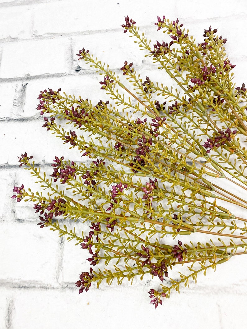 May include: A bunch of artificial greenery with small purple flowers. The stems are green and the flowers are a deep purple color. The greenery is arranged in a fan shape.