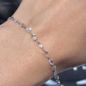 May include: Delicate gold bracelet featuring a series of clear, oval-shaped gemstones. The gemstones are linked together by a fine gold chain, creating a sparkling and elegant accessory. The bracelet is shown on a person's wrist.