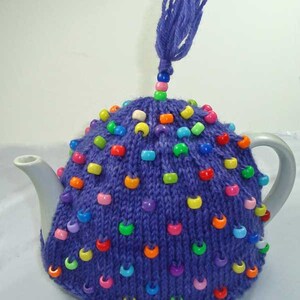 May include: A purple knitted tea cozy adorned with colorful beads. A matching purple yarn tassel is attached to the top. The cozy is designed to fit a standard teapot, providing insulation and a decorative touch.