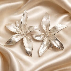 May include: A pair of silver-toned earrings shaped like open lily flowers. Each earring features five elongated, textured petals with a metallic finish that reflects light, creating a bright, polished appearance with visible fine lines on the surface.