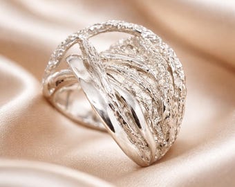 May include: A silver ring featuring a wide, open-work band composed of multiple overlapping strands. The design combines smooth, polished metallic ribbons with textured, granular sections that create a layered, dimensional appearance.