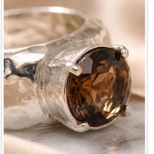 May include: A silver ring with a large, round, brown gemstone. The ring has a textured band and four prongs holding the stone. The gemstone is faceted, catching the light and displaying various shades of brown.
