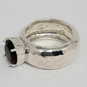 May include: A silver ring with a large, dark gemstone. The ring has a textured band and a setting that securely holds the oval-shaped stone. The gemstone appears to be a deep, rich color, possibly garnet or a similar stone.
