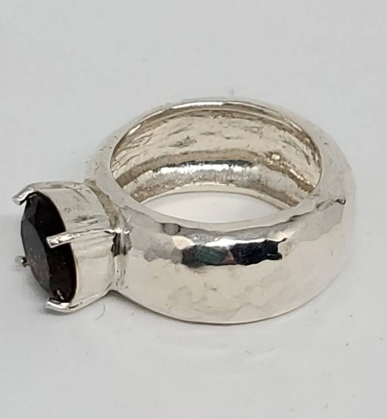 May include: A silver ring with a large, dark gemstone. The ring has a textured band and a setting that securely holds the oval-shaped stone. The gemstone appears to be a deep, rich color, possibly garnet or a similar stone.