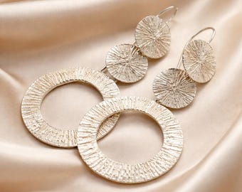May include: A pair of silver-toned drop earrings featuring a tiered design. Each earring consists of a small textured circular disc connected to a larger textured circular hoop. The metal surface has a radial, sunburst-like etched pattern.