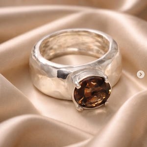 May include: A silver ring with a large, oval, brown gemstone. The ring has a thick band with a hammered texture. The gemstone is set in a four-prong setting. The ring is displayed on a satin fabric.