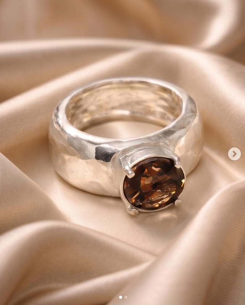 May include: A silver ring with a large, oval, brown gemstone. The ring has a thick band with a hammered texture. The gemstone is set in a four-prong setting. The ring is displayed on a satin fabric.
