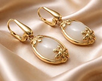 May include: A pair of gold-toned drop earrings featuring oval-shaped, smooth white stones. Each earring is accented with a small, metallic floral-shaped detail at the top and bottom of the stone, secured with a hinged lever-back closure mechanism.