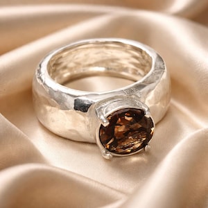 May include: A silver ring with a large, oval-cut, brown gemstone. The ring has a thick band with a hammered texture. The gemstone is set in a four-prong setting, and the ring is displayed on a satin surface.